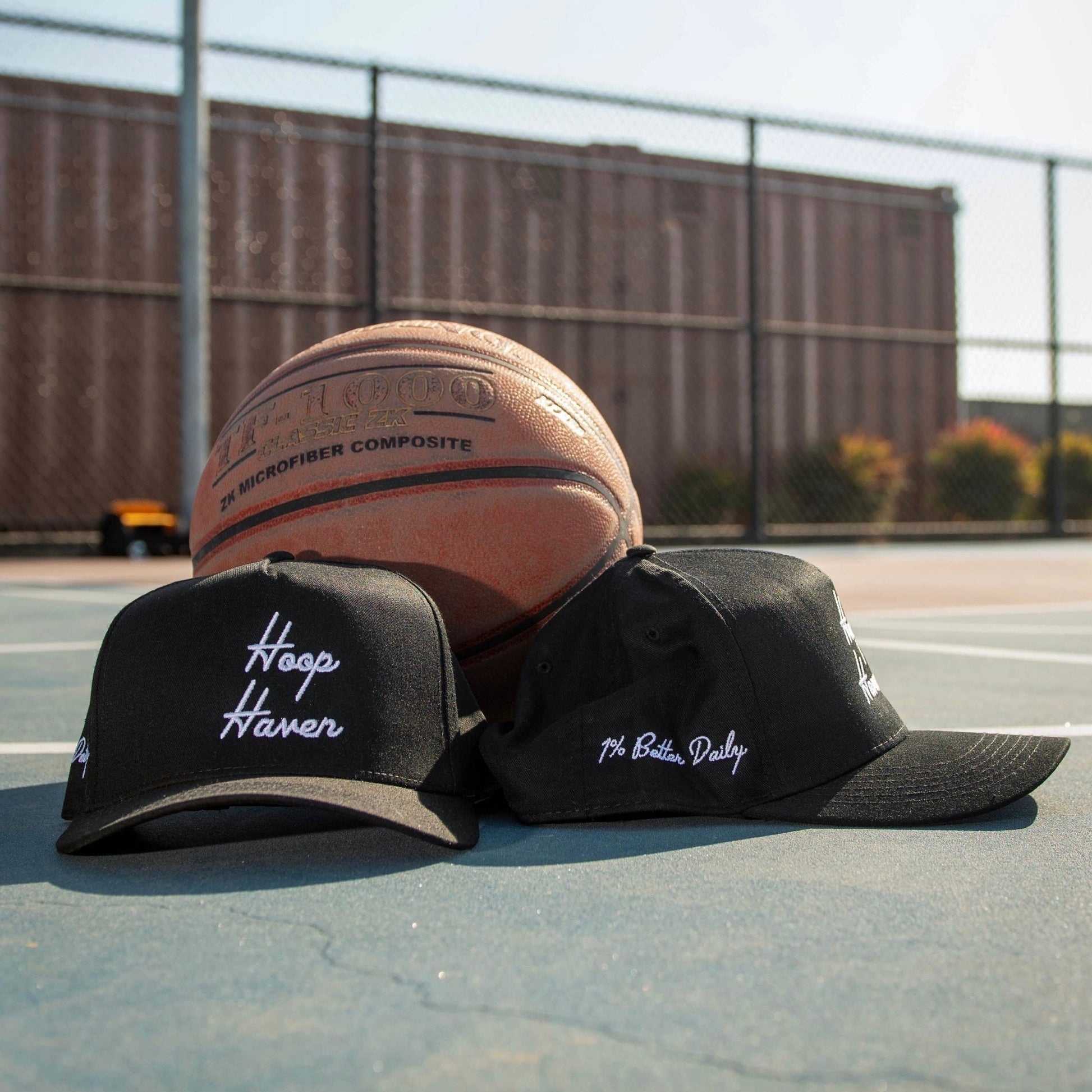 Two Black Hats on a basketball court. The hats have white text on them that say "Hoop Haven" in a cursive font. 