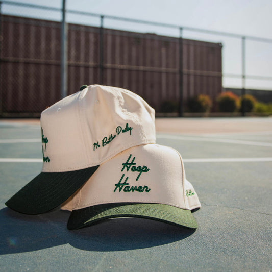 Two Cream colored hats on a basketball court. The embroidery on the hats read "Hoop Haven".